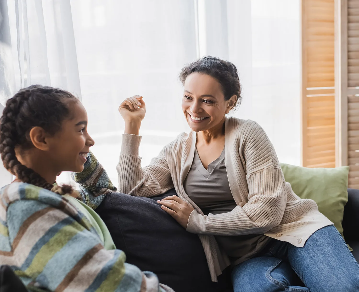 Een glimlachende volwassen vrouw in een beige vest voert een ontspannen en warm gesprek met een jonger meisje op een bank in een lichte kamer. Als onderdeel van ambulante begeleiding door Stichting Biozorg.